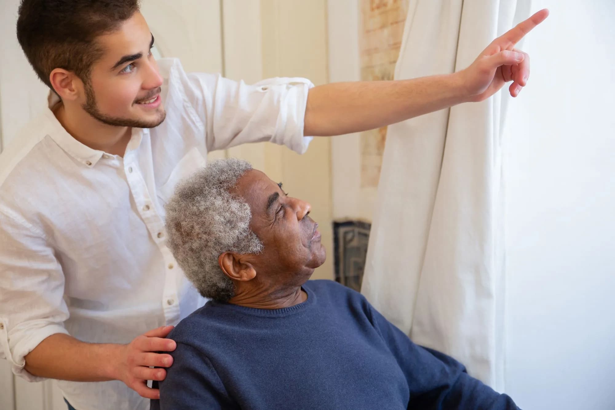 A younger man resting a hand on an older man's shoulder as they look out of a window together