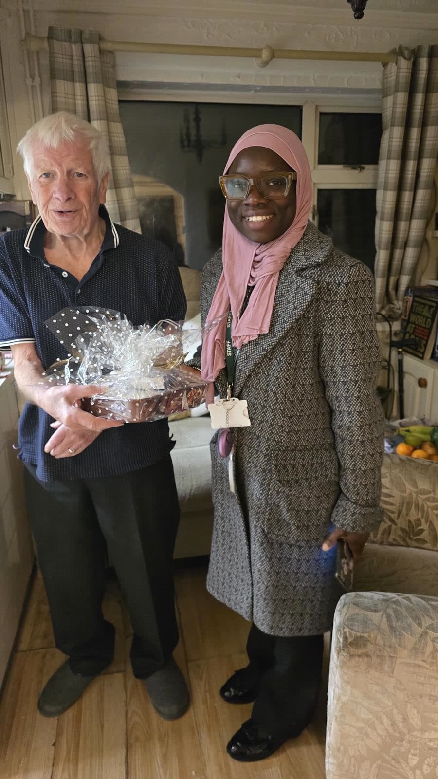 An older man holding a Christmas hamper beside a Nema staff member in his home