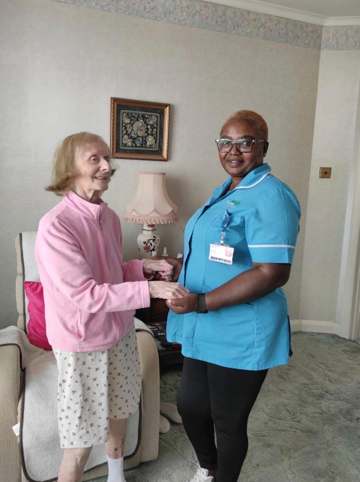 A Nema carer in a turquoise uniform holding hands with an older woman in a pink jacket, in her living room